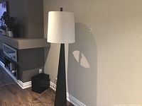 Full view of the floor lamp showing the tall black metal tapered base and the white round fabric shade.