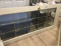 Full view of French door leaning against a wall, showing cream-colored painted wood and large bevelled glass area with brass pane separators.