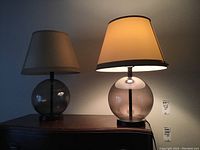 One lamp illuminated showing light beige conical fabric shade on large smoked glass spherical base beside matching unlit lamp with some piping damage to shade.