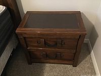 Full view of wood night table showing two drawers with leather-like handles and black top inset, against carpet and mattress.