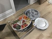 Overview photo showing the resin charger plates stacked, the boxed ceramic divided serving dish, and a stack of disposable plastic divided trays.