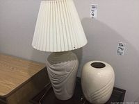 Photo showing ceramic table lamp with pleated shade next to ceramic vase, both off-white with carved swirl design.