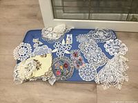 Overview of the 13 hand made doilies arranged on a blue tray, showcasing variety in size, shape and embroidery styles.