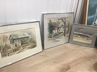 Three framed watercolour paintings displayed leaning against a wall on wood floor, showing different rural and landscape scenes
