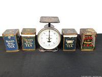 Front view of postal scale and lined up five tea tins, showing labels and scale dial.