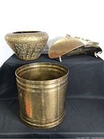 Photo showing all three brass items arranged on a black cloth: brass planter front and center, ornate pot behind left, fireplace log holder behind right.