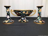 Front view of the three-piece set with the footed bowl in the center and two candle holders on sides showing black background and colorful floral hand painting with gold trim.