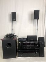 Two Denon receivers stacked center, subwoofer at left, cables coiled on top, 2 bookshelf speakers on 37" stands behind, center channel speaker atop receiver, 2 wall speakers at right