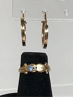 Gold ring with leaf design and small blue stone displayed on black stand, with hoop earrings hanging above.