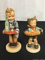 Both Hummel figurines displayed side by side on a black surface with a white background.