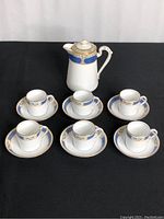 Front view of coffee pot with six demi tasse cups and saucers arranged on black surface