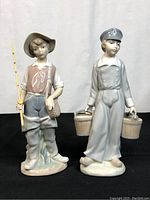 Photo of two Lladro porcelain figurines displayed on black surface: Dutch boy with two pails in light gray and brown tones and boy with fishing pole and basket, approximately 8.5 inches tall each.