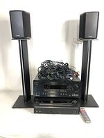 Front view showing ONKYO AV receivers stacked with tangled connection cables on top, flanked by Paradigm bookshelf speakers on stands, and remote control to right.