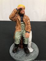 Front view of seated pilot figurine wearing yellow hat, brown coat, green overalls, holding lifebuoy