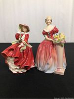 Two Royal Doulton porcelain figurines displayed side by side on a black surface, one in a red dress with hat and the other in a gown with flower pedestal