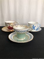 Three teacup and saucer sets displayed showing different designs and colors including mint green, pink with gold, and white with blue bird.