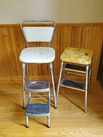 Photo showing a vintage white metal 2-step stool chair with padded seat and backrest, a yellow floral patterned small step stool with two black steps, and a small metal 2-step stool with black steps and silver legs.