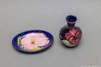 Two Moorcroft pieces: a small bud vase and an oval dish placed side by side on a gray surface, showing floral designs.