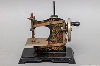 Front left side view of vintage Mueller toy metal sewing machine with decorative painted floral patterns and hand crank.