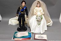 Photo of the Prince Charles and Princess Diana dolls standing side by side in wedding attire with certificates and booklet in front.