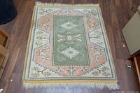 Full view of a rectangular handmade Turkish wool rug showcasing central green geometric design with peach, cream, and blue floral borders, fringed edges, laid on wood flooring.