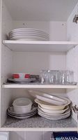 Photo of stacked white plates on top shelf, glass tumblers, mugs and bowls on middle and bottom shelves inside cabinet.