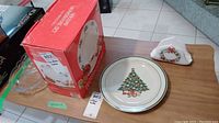 Photo showing boxed 12-piece Corelle Christmas dinnerware set, glass dish packaging, and ceramic napkin holder on wooden table.