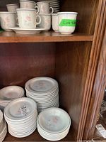 Photo showing stacked ceramic cups and saucers with floral design, alongside bowls and plates on a wooden shelf.
