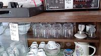 Glassware and white ceramic dishes with floral decorations arranged on wooden shelves including glasses, coffee cups, saucers, vases, and bowls.