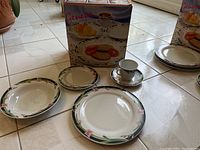 Photo showing a selection of the dinnerware set including plates, a cup and a saucer, and the box with Geneva branding.