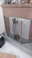 Fireplace with metal screen and metal fireplace tools set on stone hearth. Screen is three-panel with brass frame and mesh center. Tools include poker, brush, tongs, and shovel on a stand.