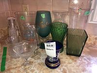 Nine vases of various glass and ceramic materials on kitchen counter, showing shapes and colors including green, blue, and clear glass and ceramic.