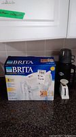 Brita replacement water filter pitcher box and black thermos with two black cups on a kitchen counter
