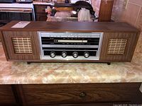 Front view of vintage stereo radio console on countertop showing woodgrain finish, speaker grilles, dial panel, and control knobs.