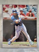An image showing Vernon Wells swinging a baseball bat in a game, signed in blue ink across the photo. Visible certification sticker for authenticity.