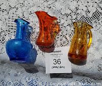 Three small crackle glass pitchers in blue, orange, and amber yellow placed on lace cloth with MaxSold tag, showing front and side views