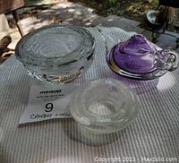 Photo showing three glass items: a large clear glass candle holder with cut swirl patterns, a small clear round candle holder, and a purple glass oil lamp with clear handle and base.
