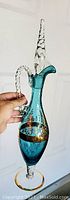 Photo showing the decanter held upright to highlight the twisted handle, elongated stopper, and gold detailing on aquamarine glass.