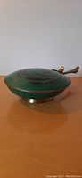 Side view of the round green enamel brass silent butler with matching spoon resting on top.