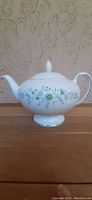 Side profile of white Wedgwood teapot with floral pattern and lid on wooden surface against textured wall.