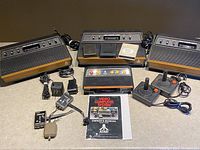 Four vintage Atari game consoles with woodgrain panels and black plastic casing, accompanied by controllers, power adapters, cartridge and manual.