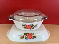 Stacked view of round JAJ Pyrex England casserole dish with clear glass lid on top of rectangular dish, both featuring pink and red rose floral patterns on white milk glass.