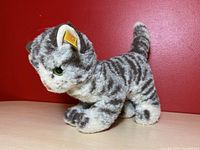 Side view of grey tabby cat plush with striped fur pattern, green eyes, and yellow Steiff tag in ear.