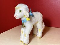 Full side view of the white Steiff lamb with yellow spots, blue ribbon, and brass bell, standing on a light wood surface against red background.