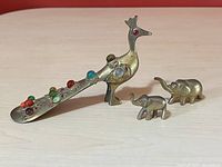 Brass peacock figurine decorated with colorful stones, facing left, with two small brass elephant figurines beside it on a light wooden surface.