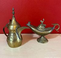 The brass oil lamp next to the brass creamer pitcher on a shelf against a red background, showing both items fully.