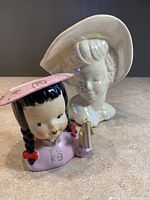 Photo of two ceramic lady head vases, one colorful little Japanese girl with pink hat and braids, other white lady bust with large hat