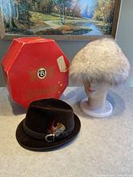 Two hats displayed with the Biltmore red octagonal box, one natural fur hat on a mannequin head and a chocolate brown velour hat with feather on a hat stand.