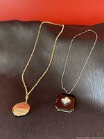 Both vintage locket necklaces displayed flat on a black textured surface showing the gold-tone circular locket necklace and the black octagonal locket with rhinestone detail.