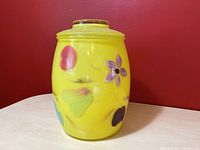 Front angle view of yellow glass cookie jar with hand painted floral decoration on a light colored surface and red wall background.